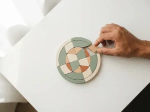 Hand plaatst houten puzzelstukje in ronde puzzel op wit bureau, zachte schaduwen, salie en crème tinten